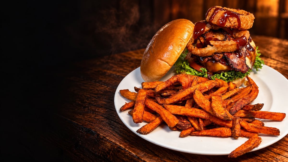 Gourmet burger stacked with bacon and lettuce on a brioche bun, served with crispy sweet potato fries on a white plate