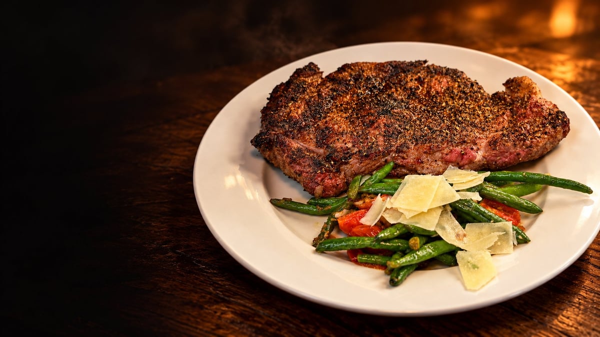 Seared steak with crusted seasoning on white plate, topped with green beans, red pepper, and shaved cheese