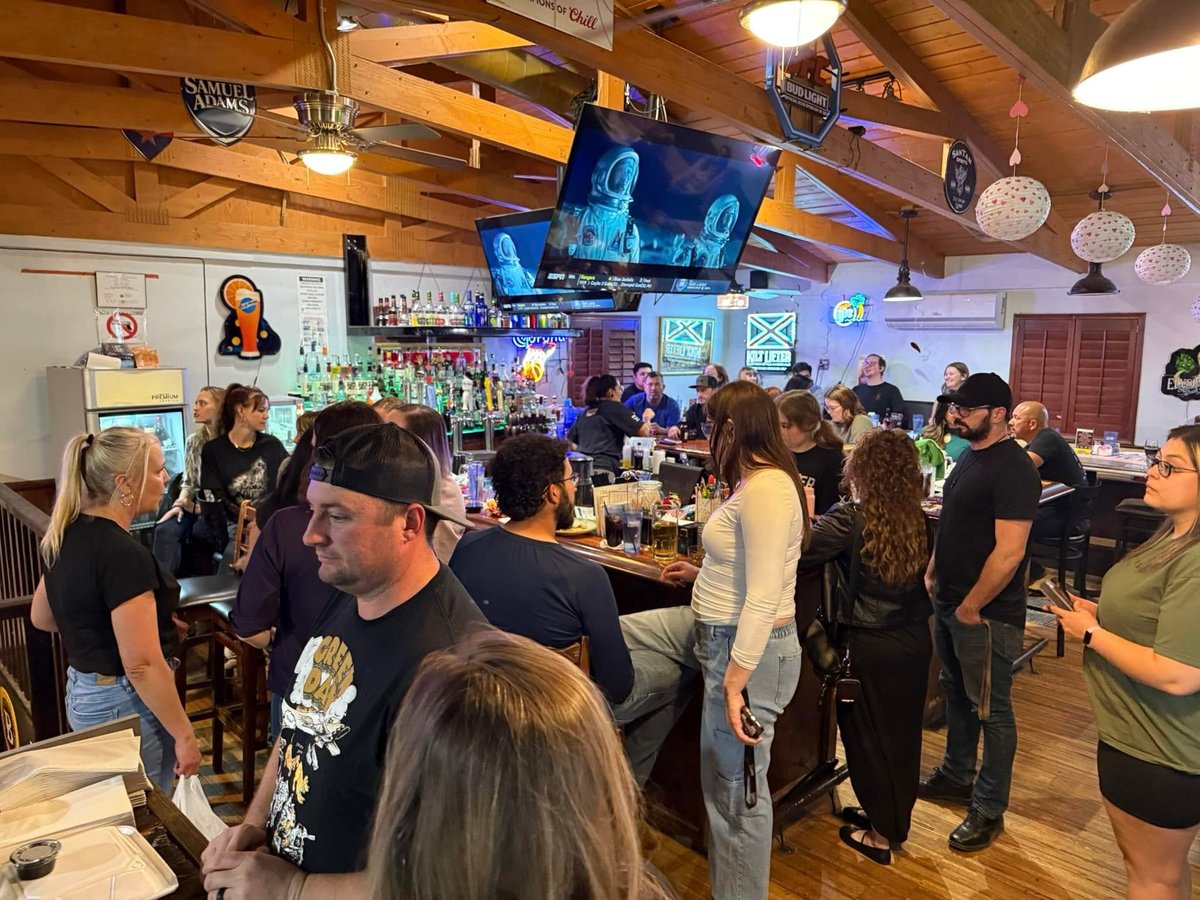 Crowded bar interior with patrons socializing, wooden beams, blue TV screens, and illuminated shelves behind the counter