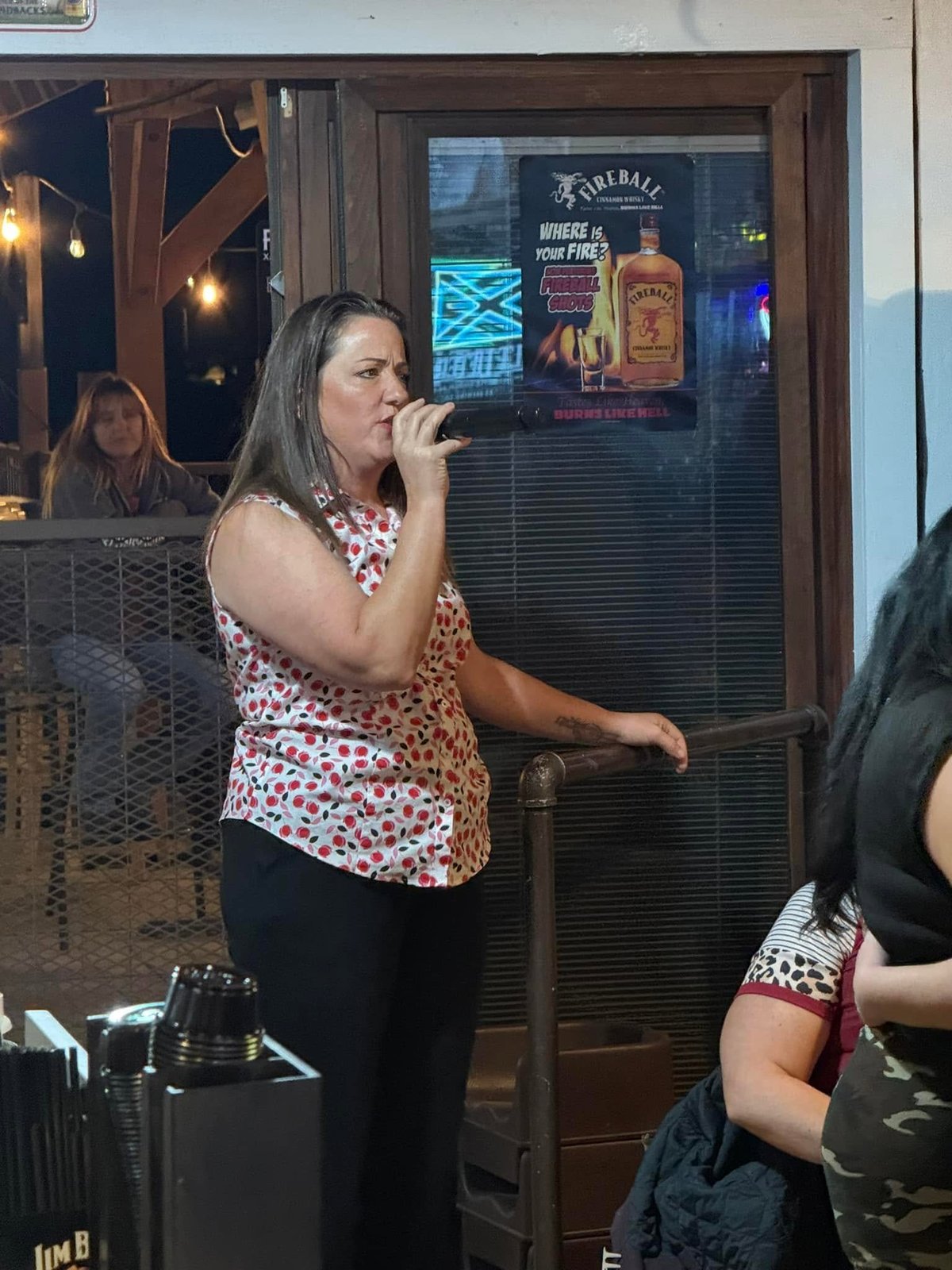 Woman singing into a microphone at a bar entrance wearing a red and white patterned top