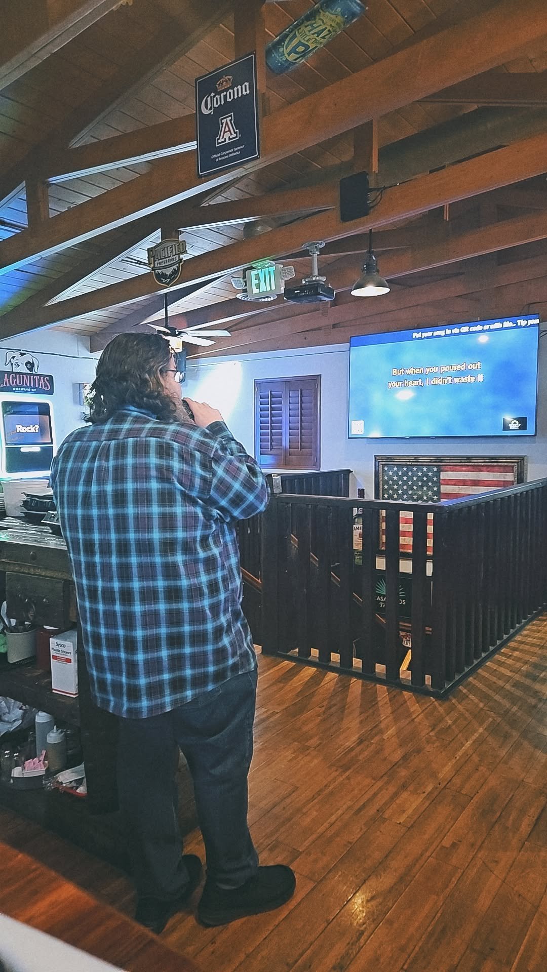 Man in a blue plaid shirt standing at a bar counter watching a TV screen with neon signs overhead
