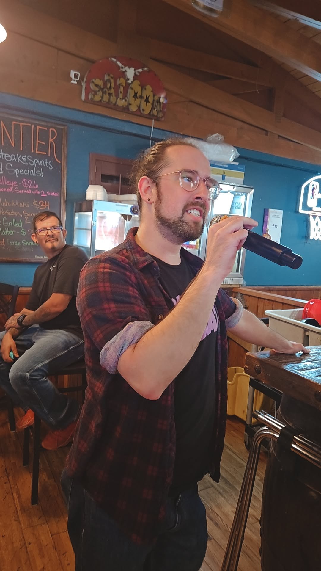 Man in plaid shirt holding a microphone speaking at an indoor venue with an audience member seated in the background