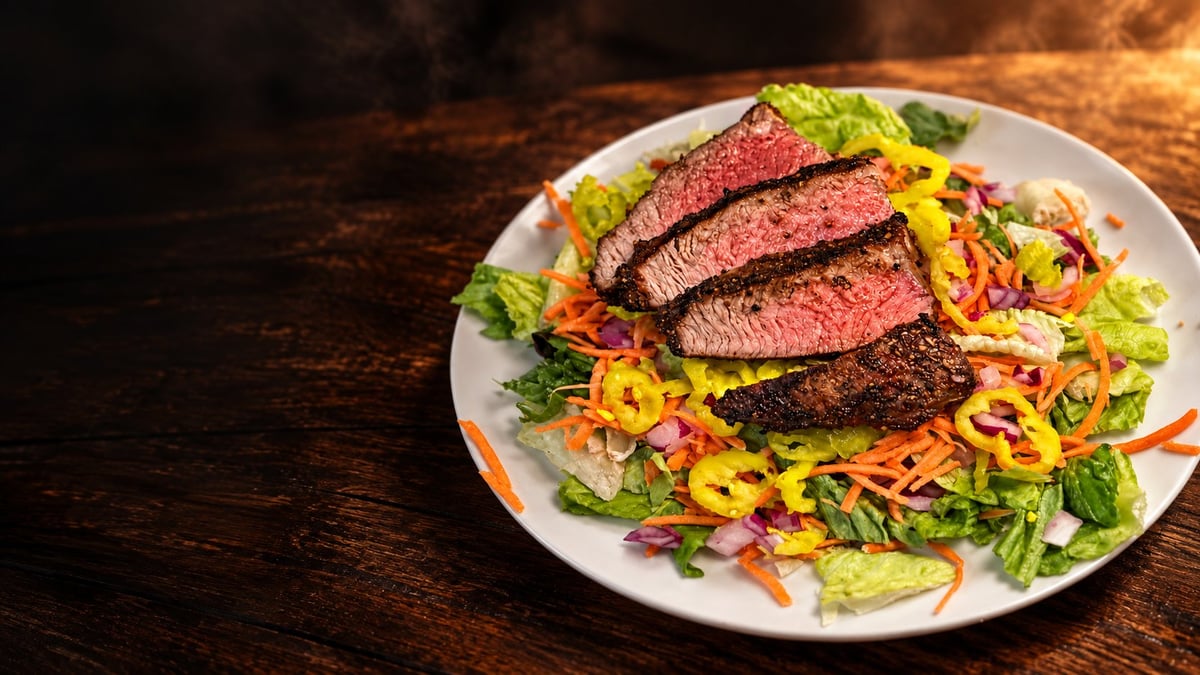 Sliced medium-rare steak over mixed salad with crispy onions and fresh greens on white plate