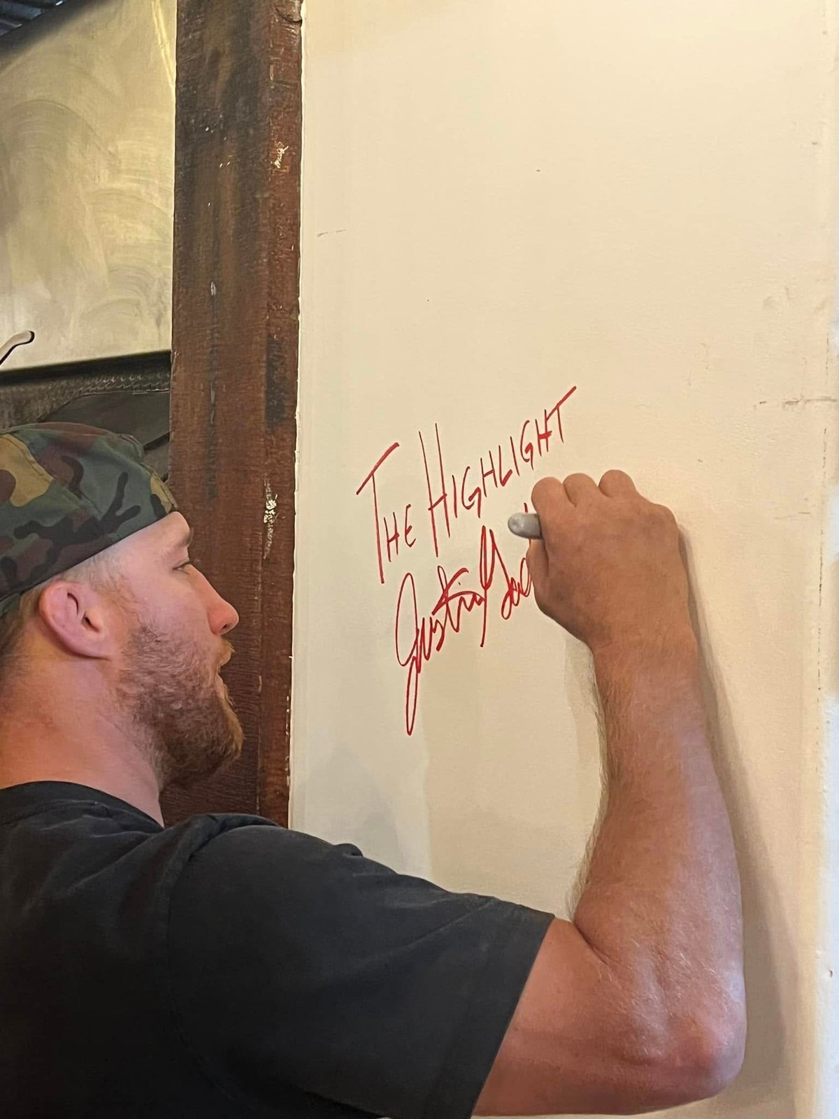 Man in a black cap writing red text on a cream-colored wall with a wooden beam visible