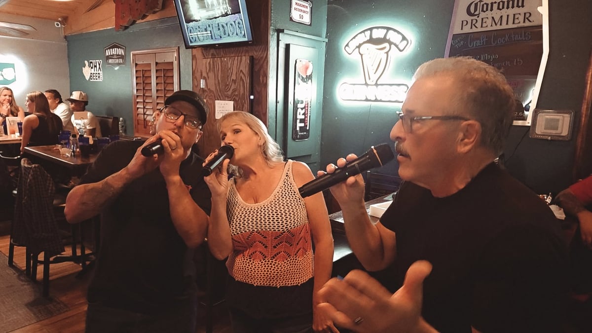 Three people singing karaoke together in a bar with neon signs and seated patrons in the background