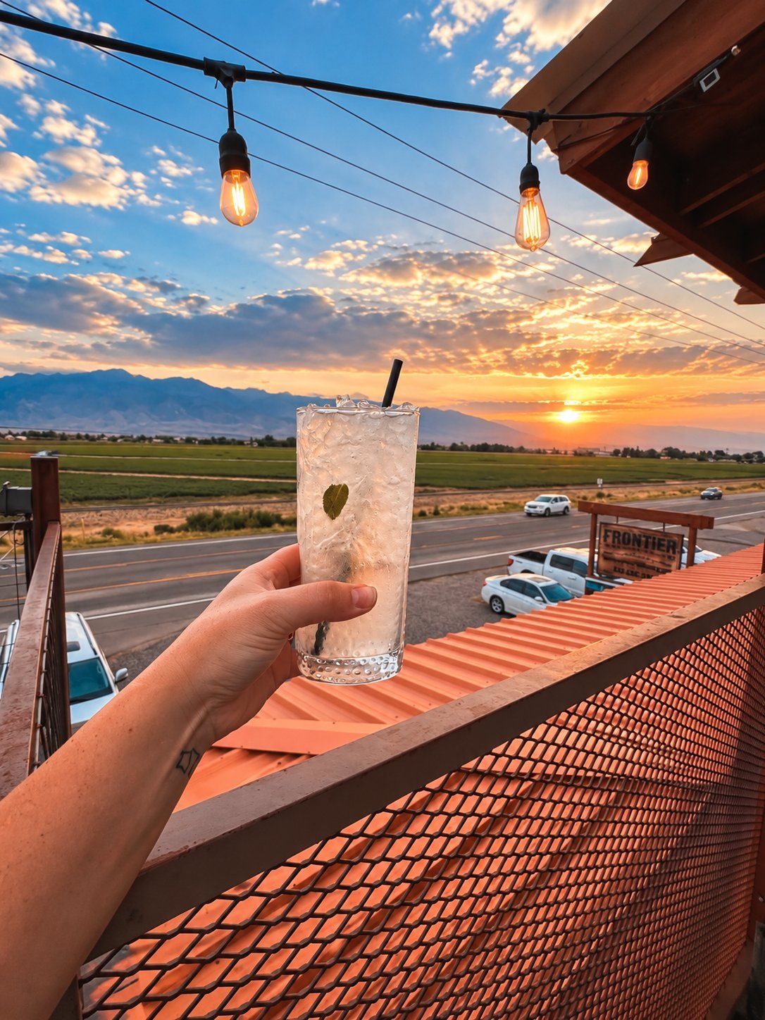 Hand holding a frosty iced cocktail on the Frontier patio at sunset with string lights and mountains in the background