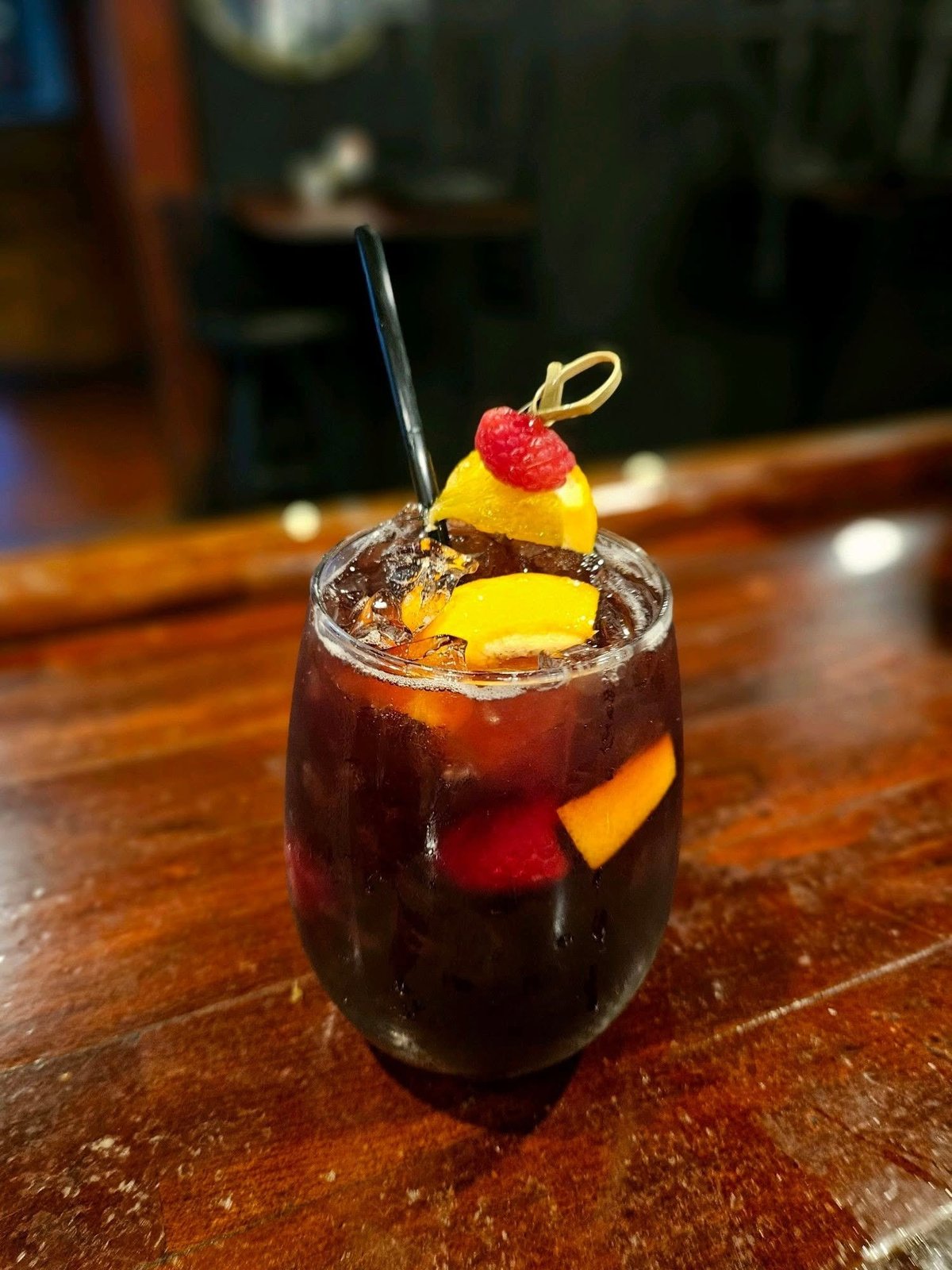 Dark cocktail with ice in a glass, garnished with yellow fruit, red berries, and a black straw on a wooden bar counter