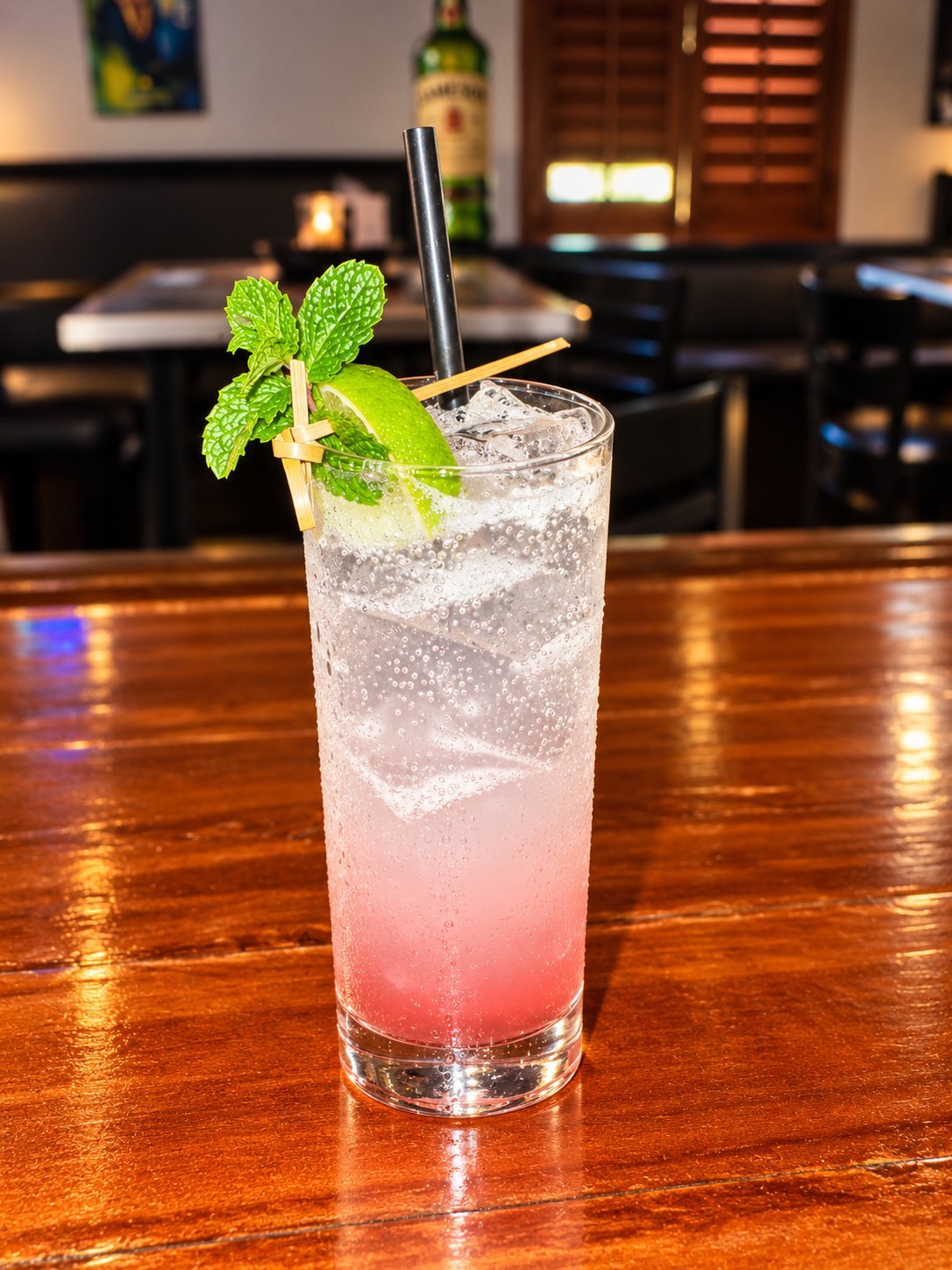 Pink cocktail with ice and mint garnish on a wooden bar counter in a dimly lit establishment