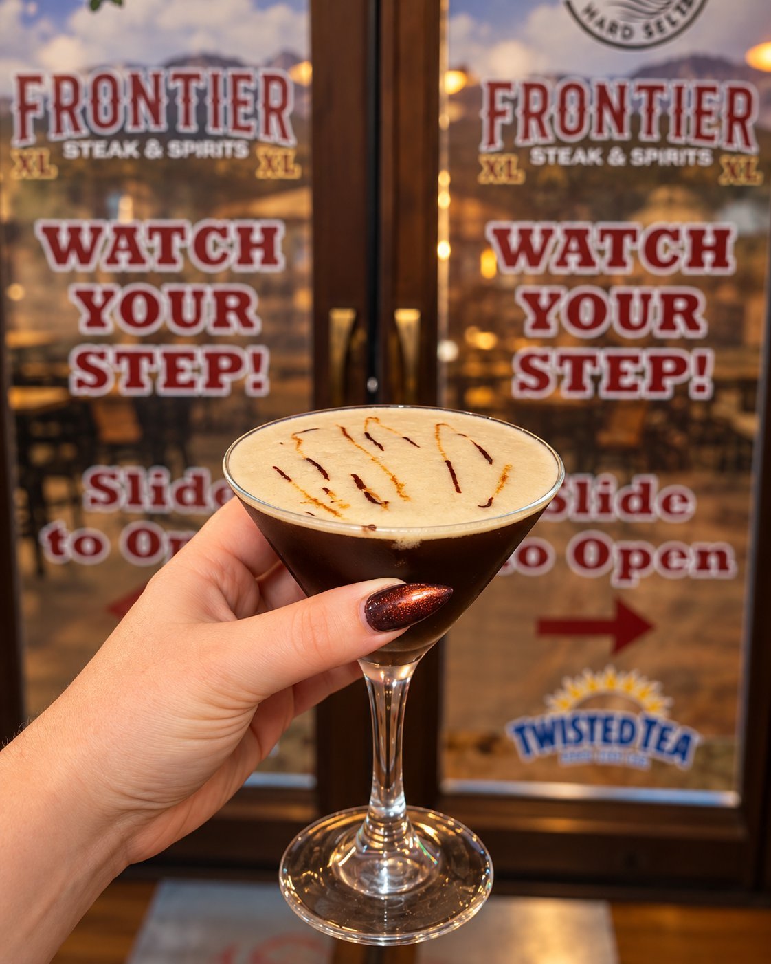 Hand holding a martini cocktail with a cream-colored drink and chocolate garnish in front of the Frontier Steak and Spirits storefront